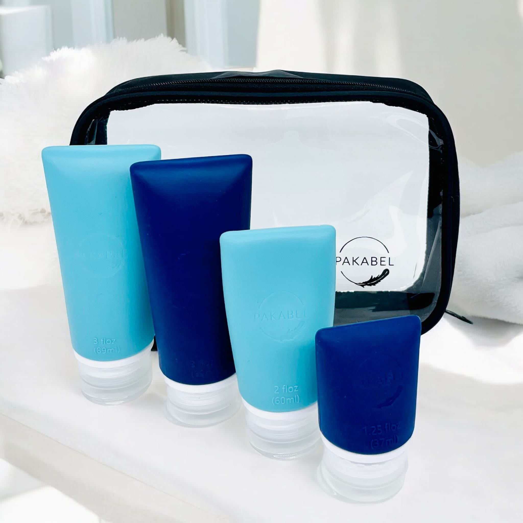 Set of 4 refillable travel bottles in three sizes and two colours. Navy blue and teal sitting in front of a clear toiletry bag on a shelf with towels in the background