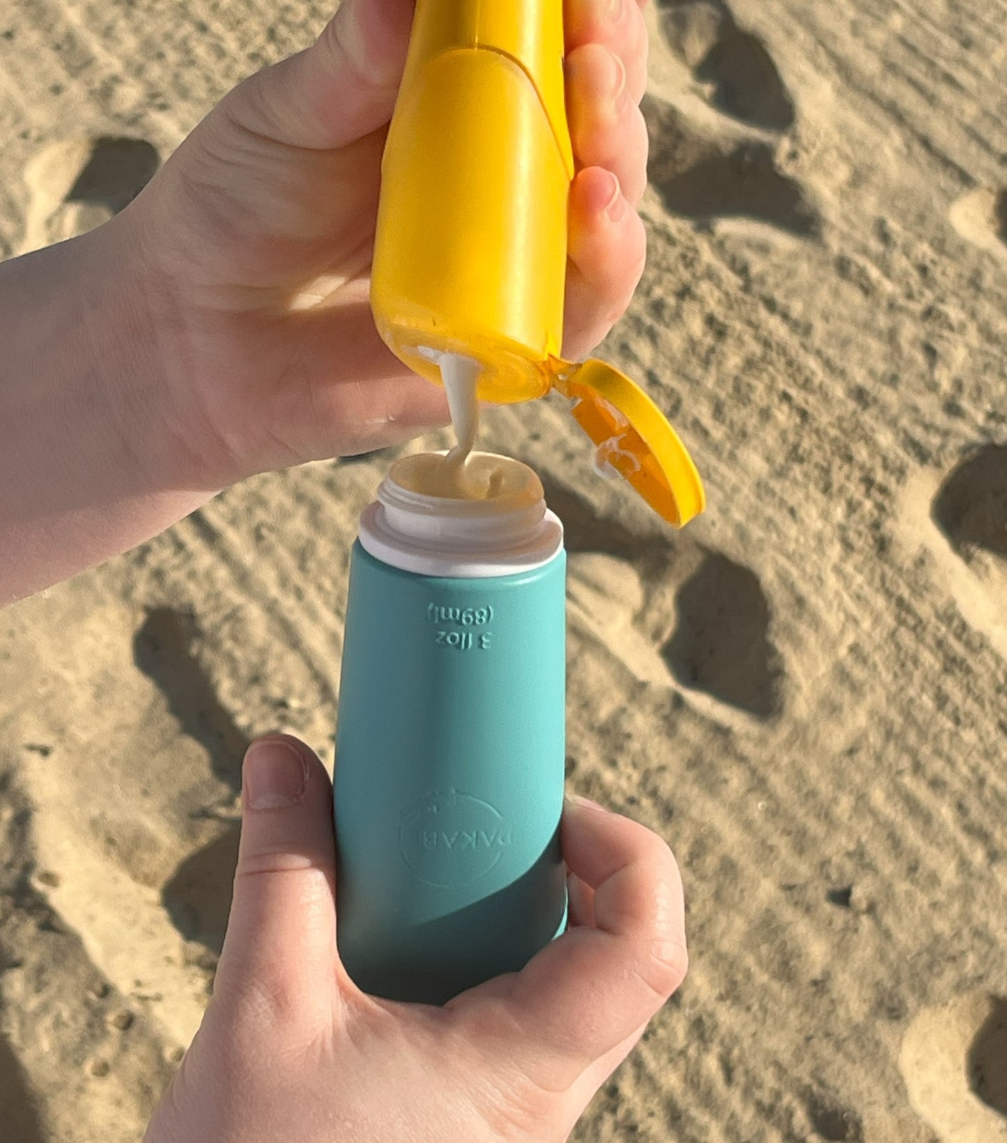 hands filling an 89 ml teal refillable container with sunblock at the beach